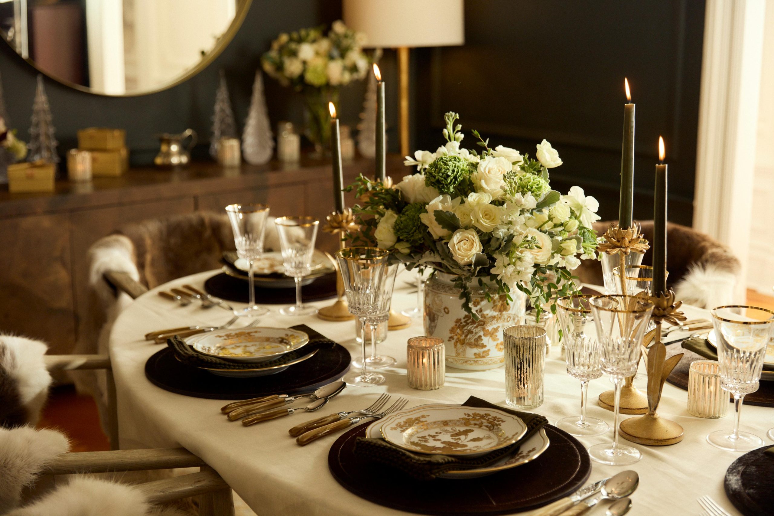 An oval dining room table set with white and gold dinnerware, a floral centerpiece, and taper candles