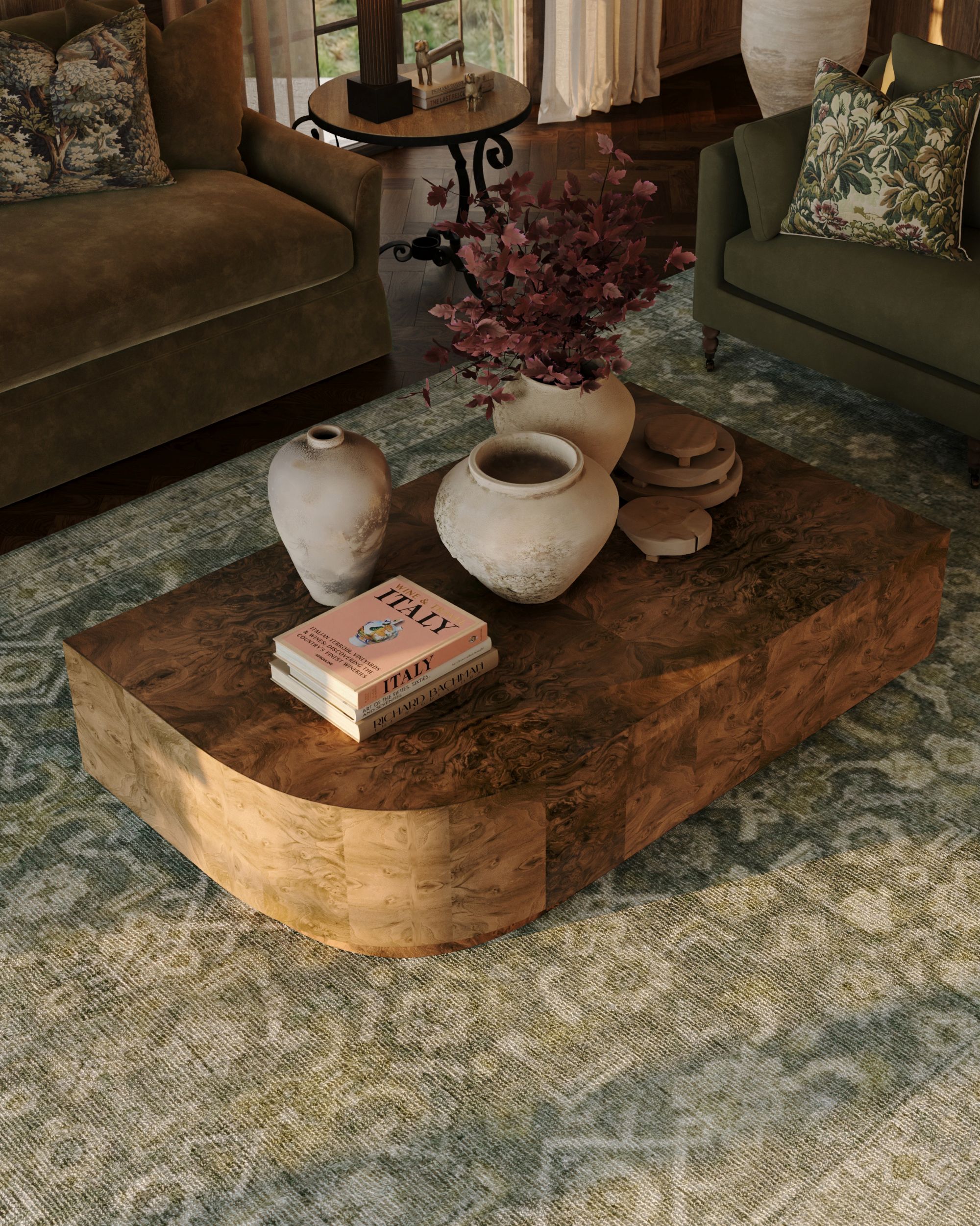 a curved burl wood coffee table topped with ceramic vases and coffee table books, in a living room with a green hand-knotted rug, green velvet sofas, and tapestry throw pillows