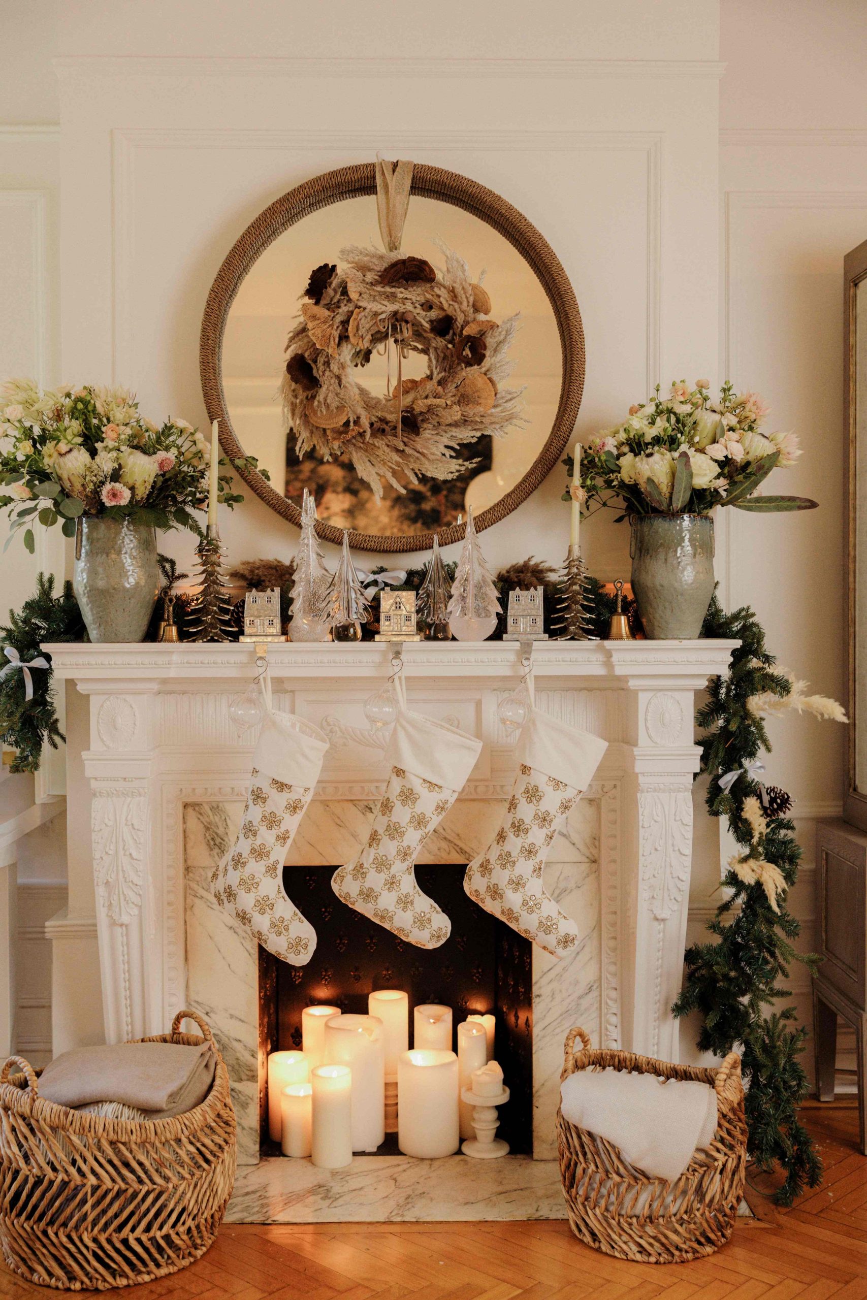 A white fireplace mantel decorated for the holidays with white and gold stockings, tapered candles, fresh flowers, a pampas grass wreath, and Simon Pierce glass Christmas trees
