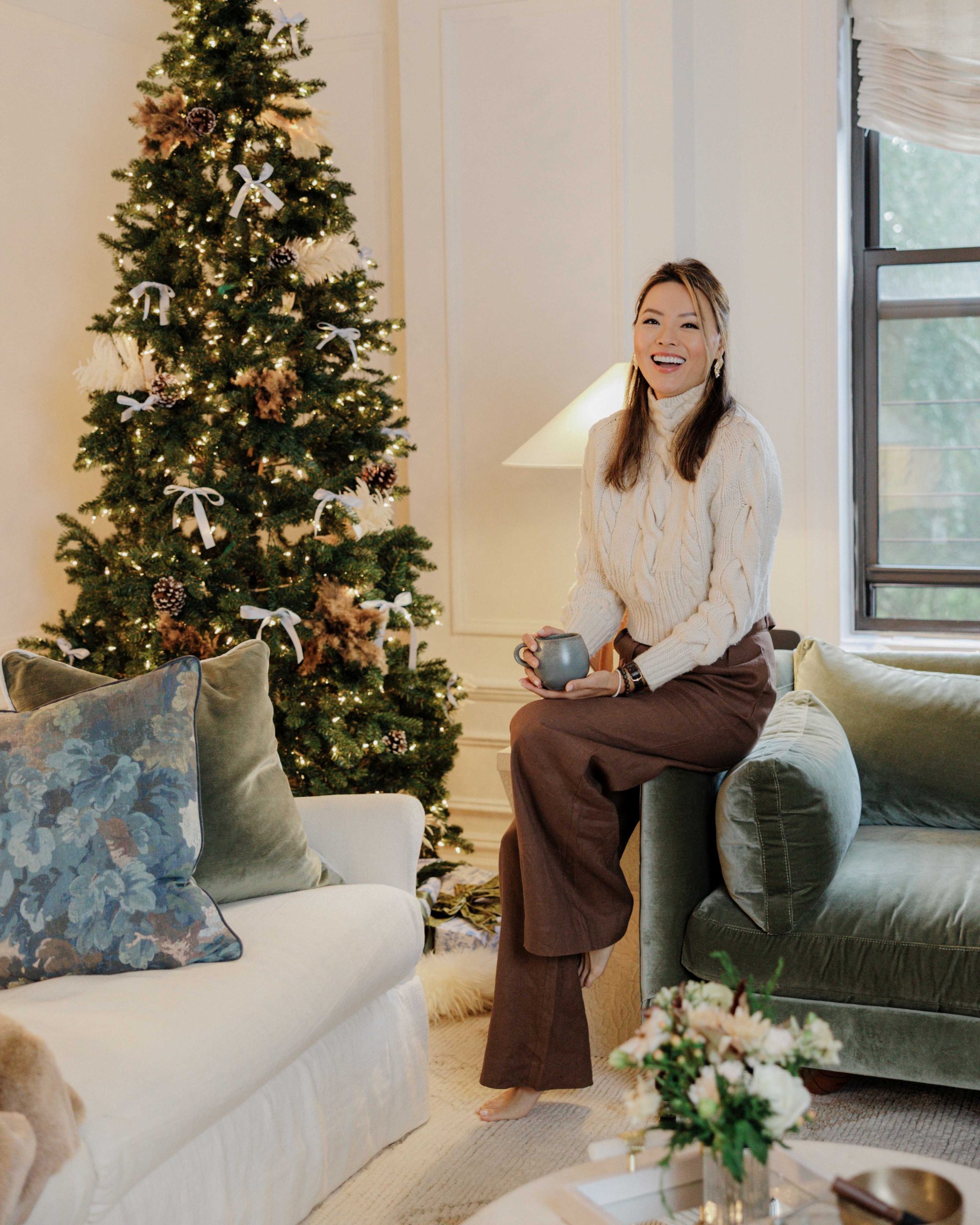 Kathy Kuo sits on the Madeline sofa in her Upper West Side apartment. The living room is decorated for the holidays with a Christmas tree, cozy throw blankets, and plush pillows