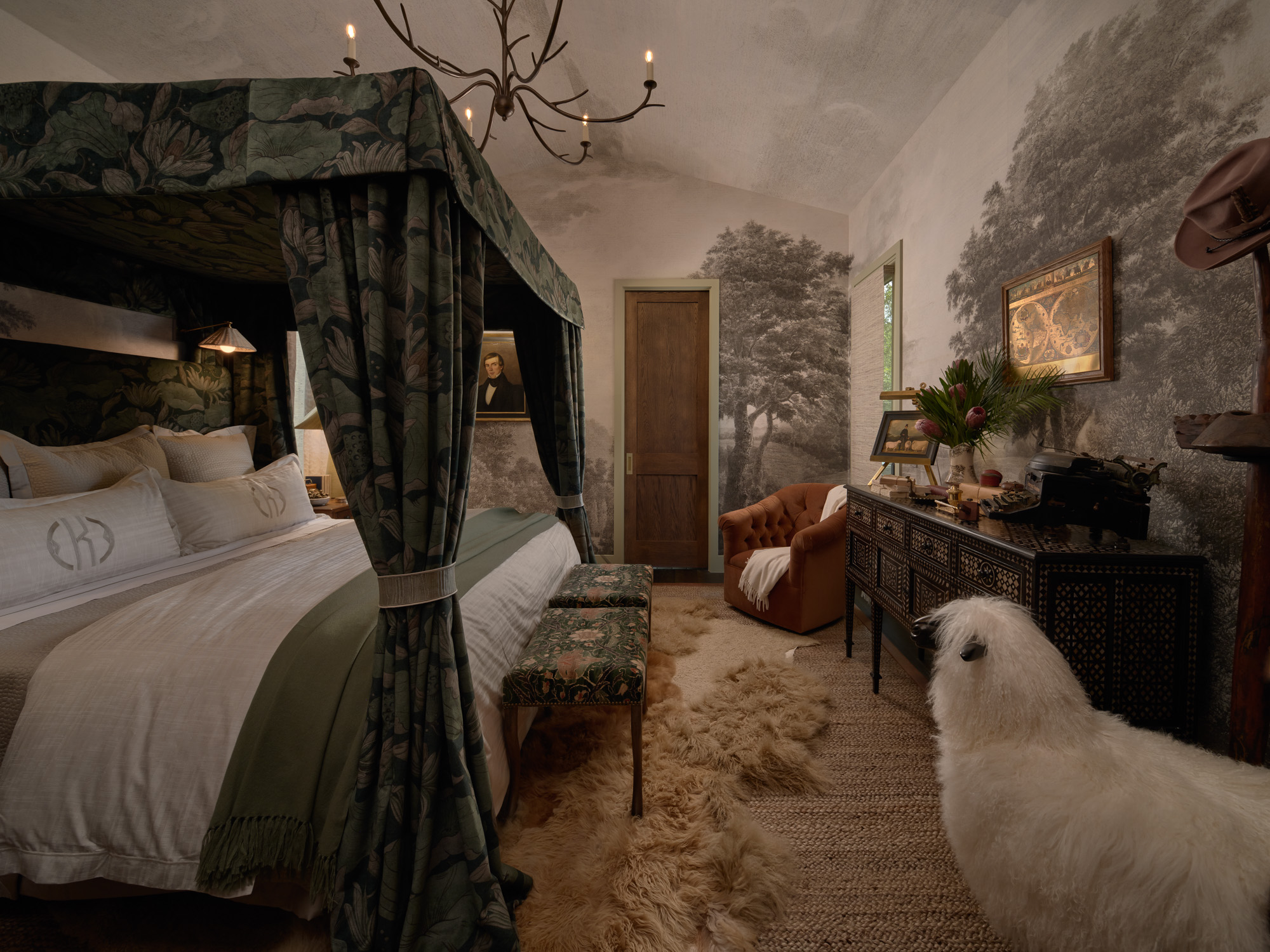 A primary bedroom with layered rugs, a four-poster canopy bed, a black sideboard with mother of pearl inlay, a bronze leaf chandelier, and a velvet swivel chair