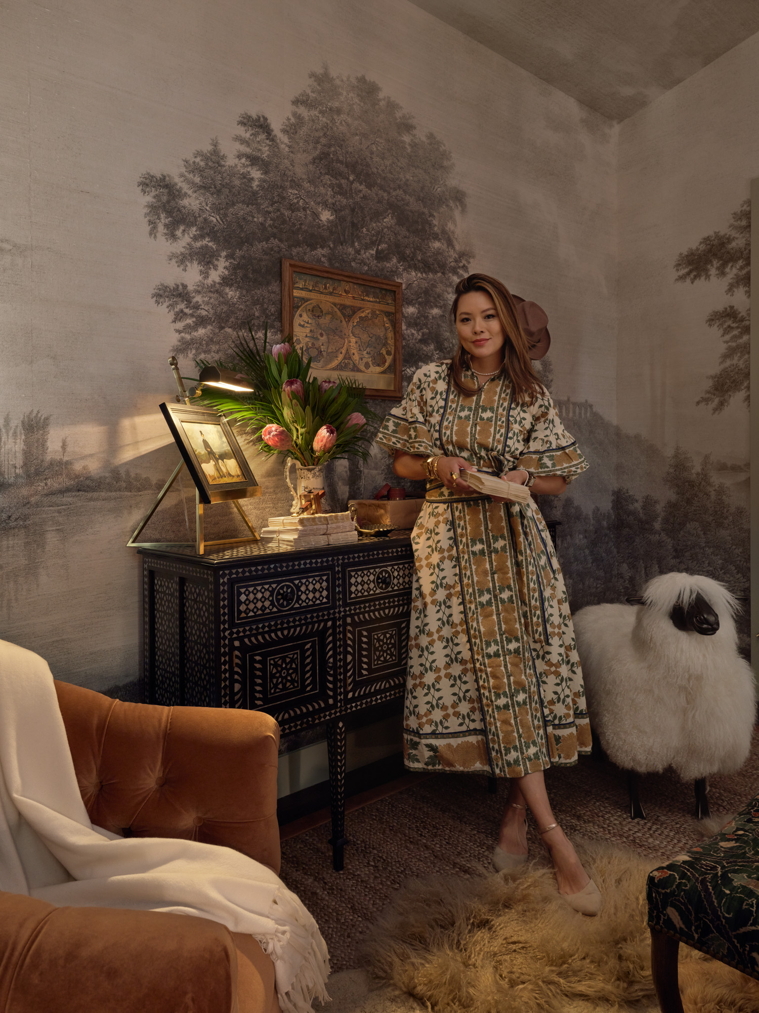 Kathy Kuo stands in a cozy bedroom with pastoral wallpaper, layered rugs, a plush velvet swivel chair, and a wood sideboard with mother of pearl inlay