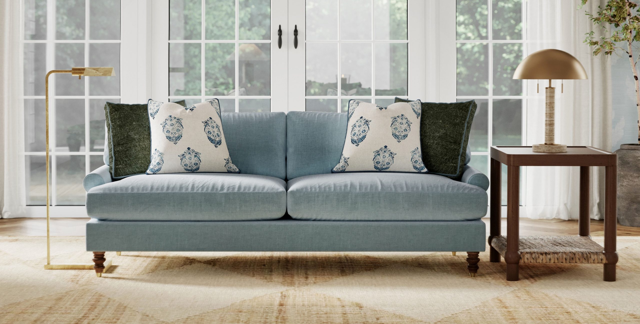 A comfortable blue velvet sofa is topped with plush throw pillows, with a gold floor lamp on one side and a side table and brass table lamp on the other