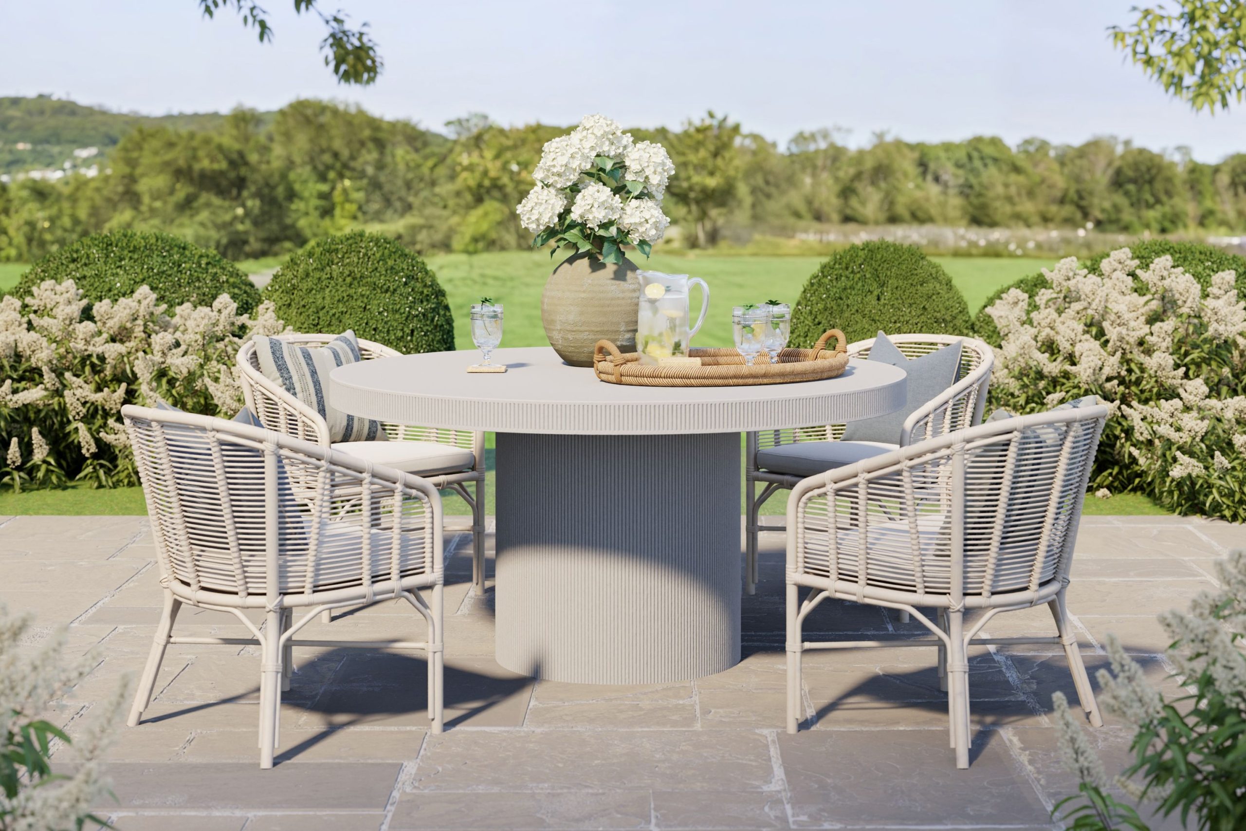 a round outdoor dining table with woven outdoor dining chairs sits on a stone patio next to an open lawn
