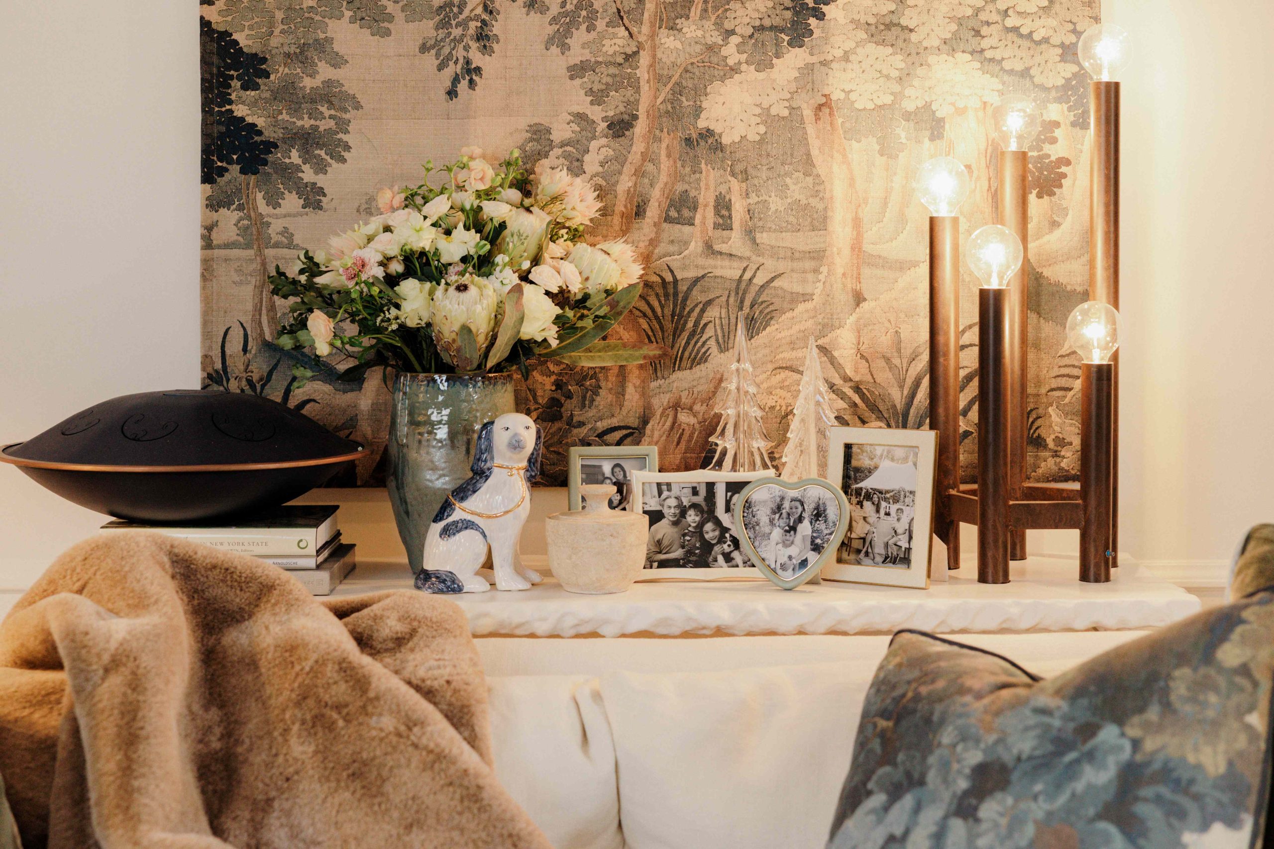 A stone console table is styled behind a sofa in a living room, with a large vase, photo frames, a decorative light, and design books on top of it