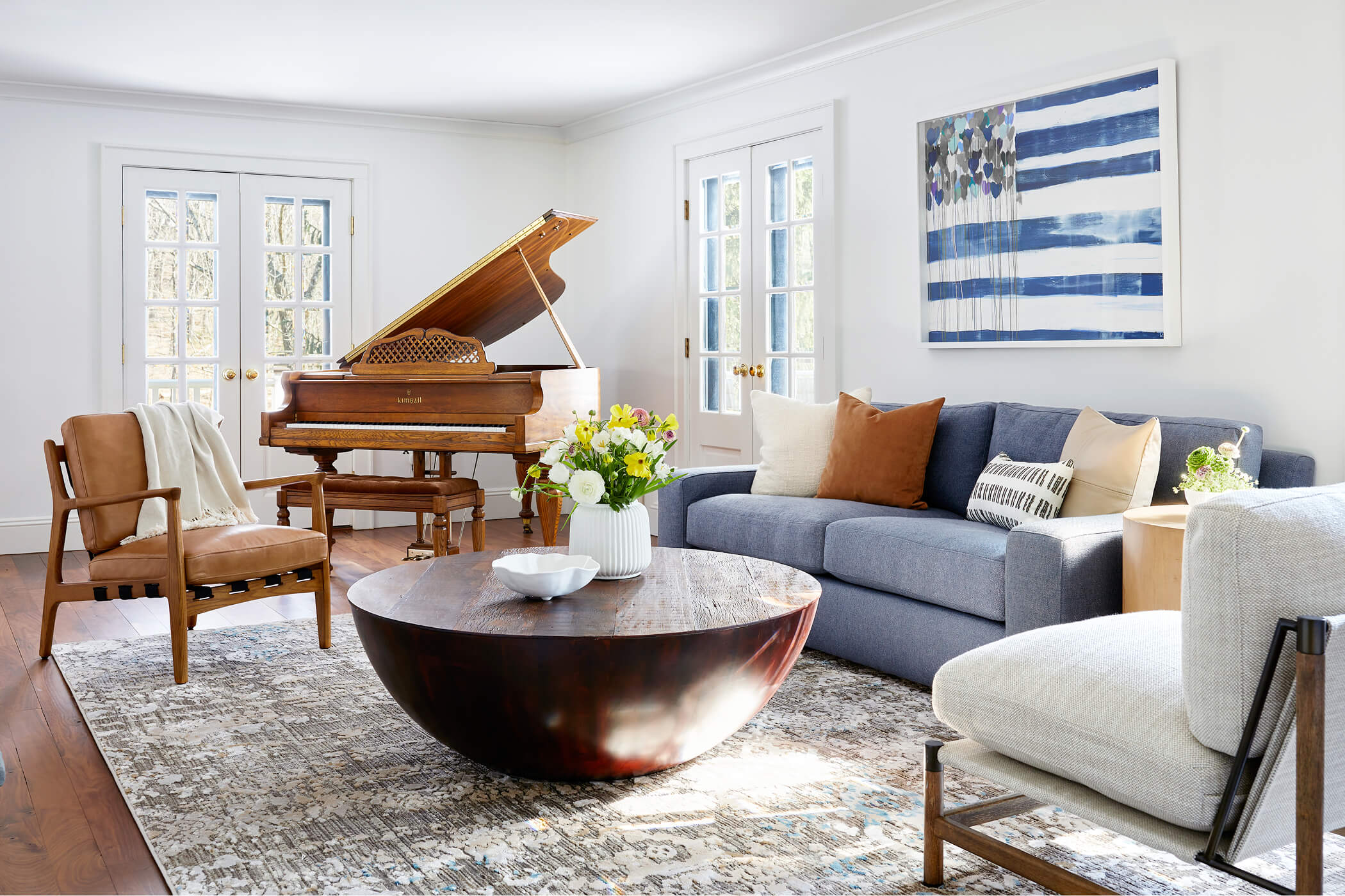 A white living room with a brown grand piano, blue sofa, round wood coffee table, and blue American flag artwork 